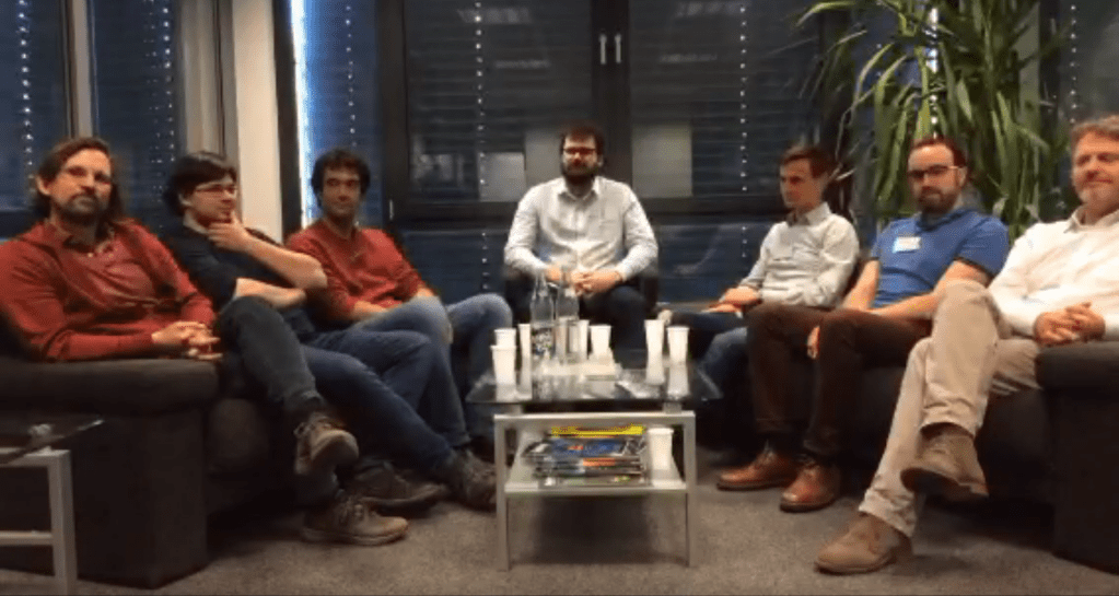 Panel of seven men sat on sofas around a coffee table.